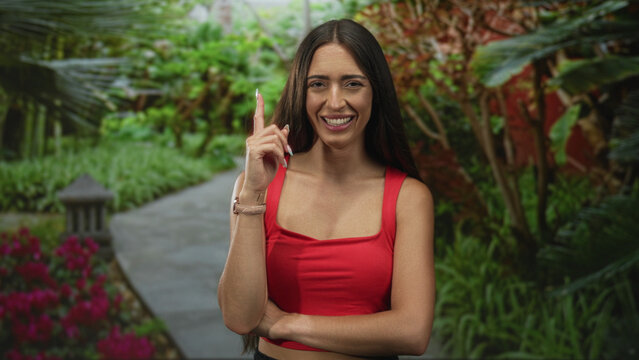 Woman points finger upward with crossed arm on park path framed by green plants and pink flowers, smiling; confidence persuasion.