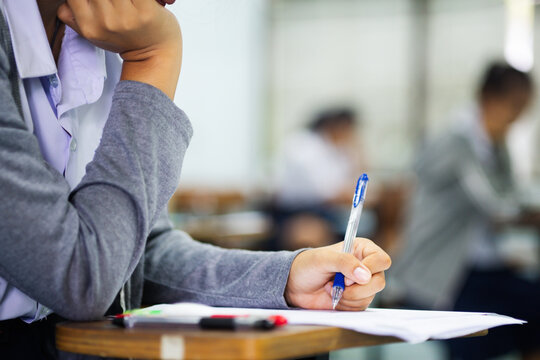 Close-up of a student's hand diligently writing on an exam paper during a school test. 
The student is using a professional pen , sitting at a wooden desk in a brightly lit classroom.