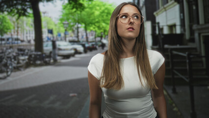 Fototapeta premium Young woman with glasses looking up while standing on a street beside parked bicycles and a building, wearing a white shirt and long hair; contemplation.