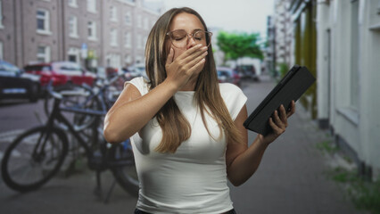Woman holding tablet and covering her mouth while reading on a city street, reacting to onscreen content  surprised wonder. © Krakenimages.com