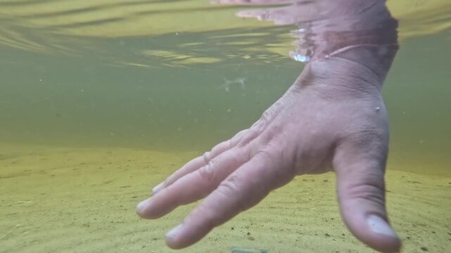 A human hand and forearm move slowly underwater in green-tinted river water, fingers spread open, then curl and shift position, floating and gesturing through clear shallow water in daylight