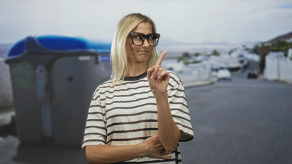 Fototapeta premium Woman wearing striped shirt and glasses points index finger up beside a large trash bin on a residential street; warning instruction.