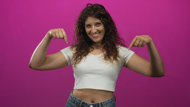Woman pointing both index fingers down at her exposed navel in a magenta studio, wearing a white cropped shirt and jeans; playful confidence.