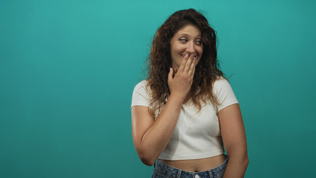 Woman covering mouth with hand and smiling in teal studio, wearing white crop top and denim jeans; playful shyness.