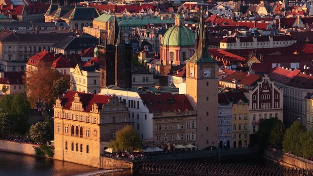 Aerial drone view of the old town riverfront with a baroque green-domed tower and clock tower above historic buildings at golden sunset.