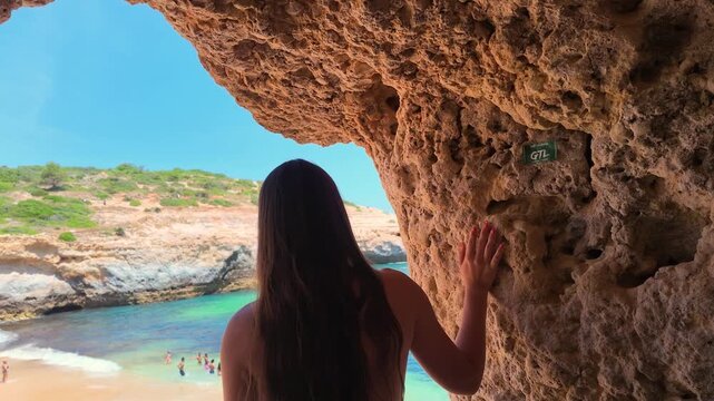Adventurous young woman walking through a rock cave and emerging to a stunning view of a hidden sandy beach with turquoise water, enjoying a sunny summer day during her vacation