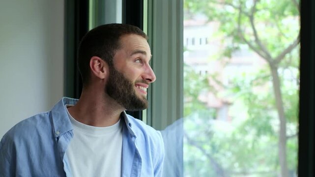Cheerful young bearded man standing at home looking out the window, feeling hopeful and optimistic while contemplating the future. His expression conveys a sense of serene happiness and contentment