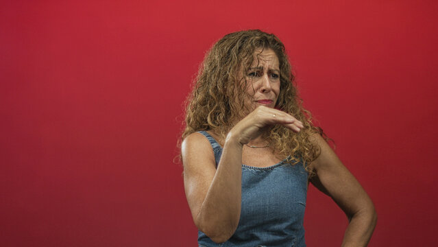 Woman pinches nose with hand in studio red backdrop wearing a denim sleeveless top and grimacing; disgust aversion smell.