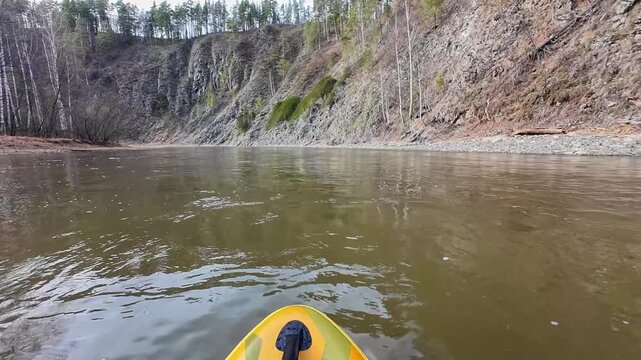 Vibrant vessel nears steep cliffs. Beng color craft approaches high rocky formations. Bright yellow kayak moves towards imposing rocky cliffs with gentle water and sparse trees