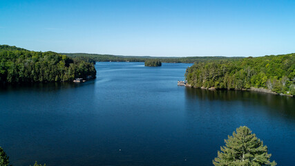 Obraz premium High-angle shot capturing a small, tree-covered island sitting in the middle of a deep blue lake, surrounded by expansive shorelines with cottages, docks and thick forest during a sunny summer day.