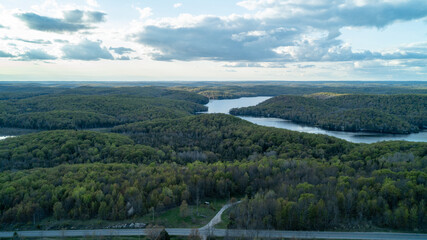 A beautiful high-angle drone photograph looking across a winding rural road towards Esson Lake, surrounded by the lush green rolling forest canopy of Tory Hill in Haliburton County during spring. © Matt