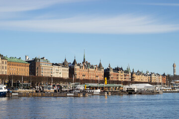 Fototapeta premium Historic waterfront buildings along the harbor in Stockholm, Sweden, featuring colorful facades, spires, and boats docked at the pier under a clear blue sky