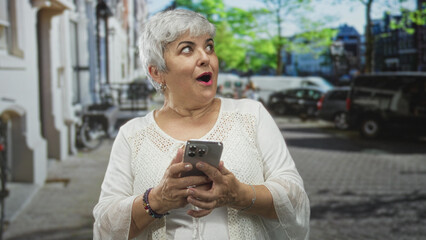 Fototapeta premium Senior woman holding smartphone, hands tapping screen on a city street while smiling and reading messages from phone; surprise connection.