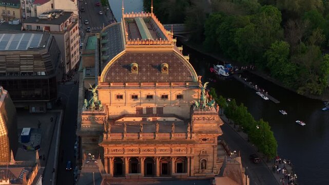 Aerial drone view directly above the ornate rooftop of the National Theatre beside the Vltava canal in golden hour light.