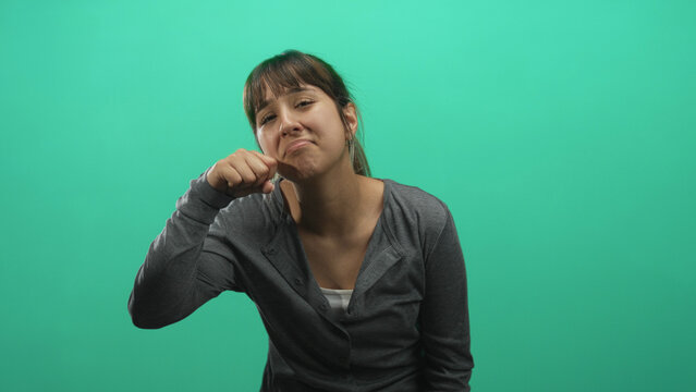 Woman in grey cardigan with long ponytail pinches her cheeks with fingers and makes a pouty smirk against a green screen in a studio; playful mischief.