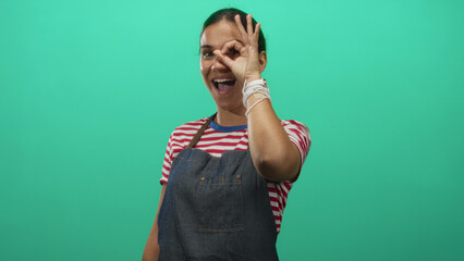 Fototapeta premium Woman making ok sign over her eye wearing denim apron and striped shirt in studio with teal backdrop; playful curiosity.