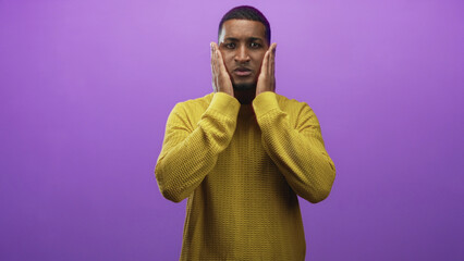 Obraz premium Man pressing hands to face in purple studio wearing yellow sweater and showing tense expression; surprise.