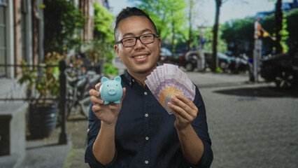 Fototapeta premium Man smiling holding blue piggybank and fanning indonesian rupiah banknotes with both hands on street; confidence savings.