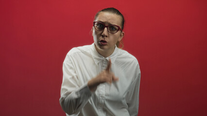 Obraz premium Woman with red glasses pointing finger and speaking, white button shirt, neutral puzzled expression against a red studio wall; uncertainty inquiry.