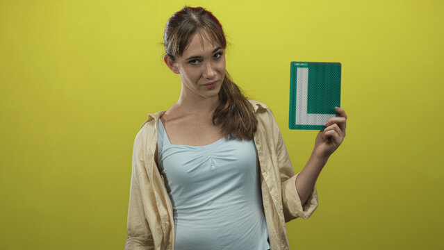 Young woman holding green learner l plate with hand to mouth gesture in yellow studio; playful learning success.