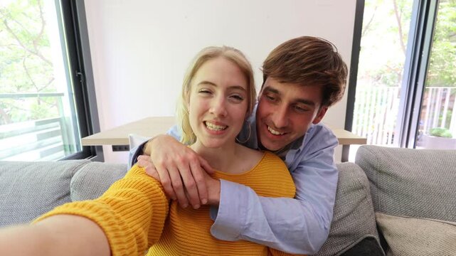 Cheerful beautiful young couple sitting on a sofa while taking a selfie video together, making funny faces and laughing in their modern apartment living room, enjoying their free time
