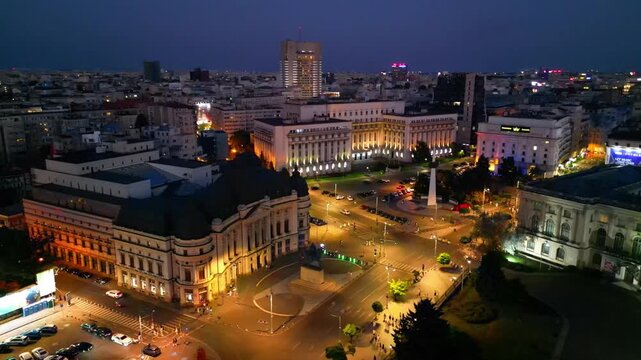 Cinematic aerial drone shot of Revolution Square in Bucharest, Romania at night. Beautiful city lights, heavy street traffic, and historic architectural landmarks under the night sky.