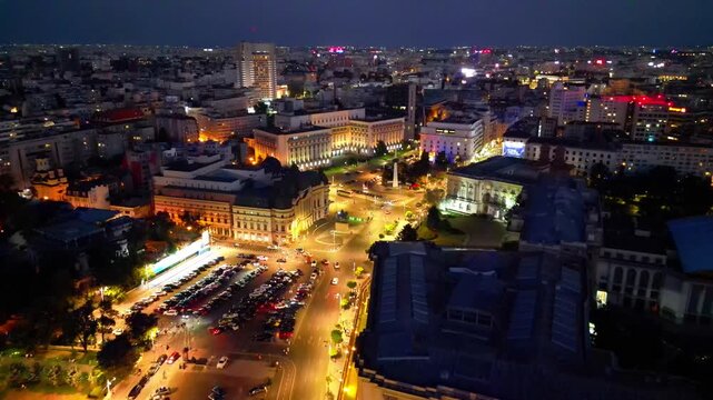 Cinematic aerial drone shot of Revolution Square in Bucharest, Romania at night. Beautiful city lights, heavy street traffic, and historic architectural landmarks under the night sky.