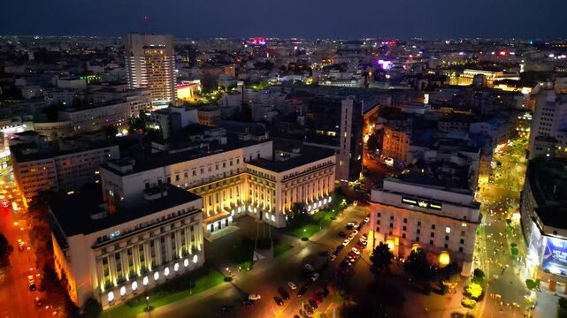 Cinematic aerial drone shot of Revolution Square in Bucharest, Romania at night. Beautiful city lights, heavy street traffic, and historic architectural landmarks under the night sky.