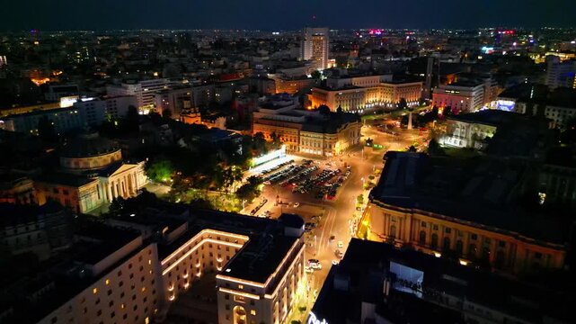 Cinematic aerial drone shot of Revolution Square in Bucharest, Romania at night. Beautiful city lights, heavy street traffic, and historic architectural landmarks under the night sky.