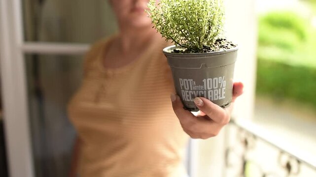 A woman holds out a grey plastic pot with thyme on a balcony. The pot features an inscription that says "pot 100% made of recyclable material".