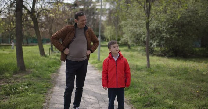 A man and a boy take a leisurely walk through a lush green park on a sunny day. They share lighthearted conversation and enjoy their time outdoors together