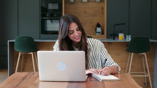 Beautiful businesswoman working from home, using a laptop and writing in a notebook while sitting at a wooden table in a modern kitchen, learning through a remote online course