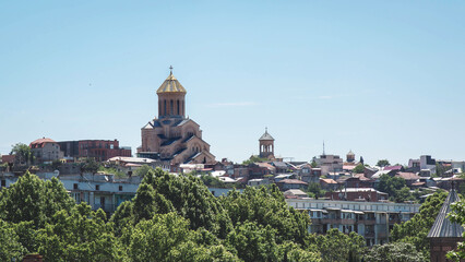 Fototapeta premium Historic skyline of Tbilisi featuring Holy Trinity Cathedral Sameba with golden dome rising above urban district, surrounded by lush green trees under clear blue sky, reflecting faith, heritage, 