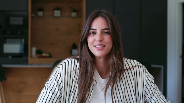Young beautiful woman having fun and puffing up her cheeks with air, making funny faces and then smiling while looking away, sitting in her modern apartment kitchen