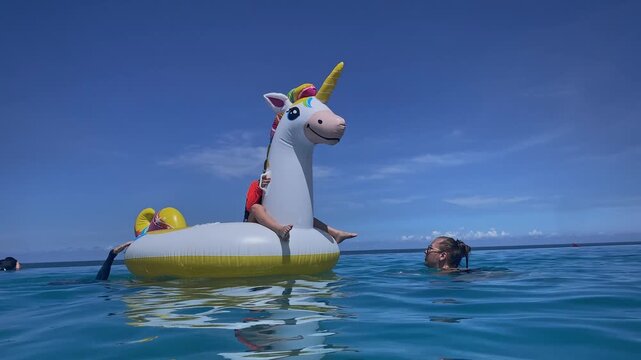 Mother and two boys on inflatable unicorn floating in pool under bright blue sky, playful splashes, visible life vest safety, calm water reflections, summer vibes and family bonding, Bali island