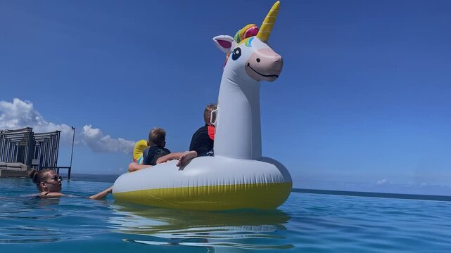 Mother and two boys on inflatable unicorn floating in pool under bright blue sky, playful splashes, visible life vest safety, calm water reflections, summer vibes and family bonding, Bali island