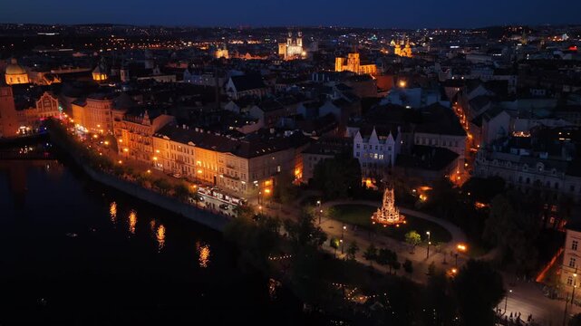 Aerial drone view of the Old Town riverfront at night with illuminated buildings, a riverside monument and Tyn Church spires visible in the background.