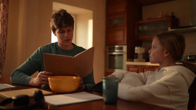 A boy sits with a girl in a kitchen. They share a bowl of food while he reads to her from a book in the afternoon