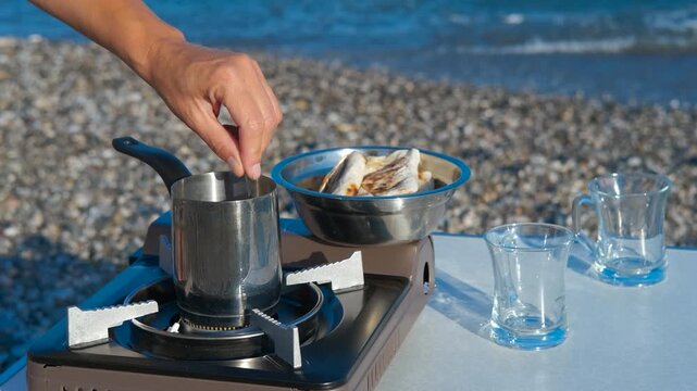A tourist makes coffee on the seashore in summer. Woman preparing morning coffee at scenic beach, adding sugar to brewing pot on portable camping stove with water glasses, flatbread, and bowl nearby