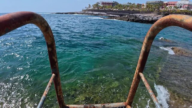 Immersive first-person perspective entering the refreshing turquoise ocean from a rusty metal ladder, revealing the beautiful rocky coastline of a sunny tropical resort on a summer day