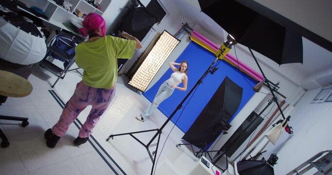 Female Photographer with Pink Hair Directs a Photoshoot with a Female Model in a Studio with a Blue Backdrop