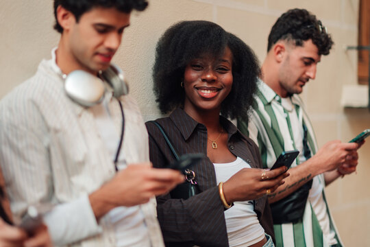Young smiling woman looking at camera while friends looking at their smartphones on city street
