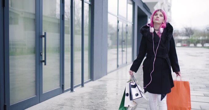 Girl with pink hair carrying shopping bags in urban setting while listening to music during a rainy day