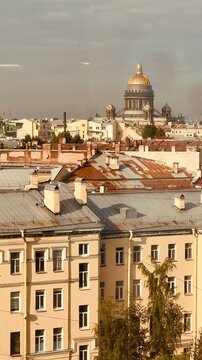Wide elevated cityscape at dusk featuring pale tiled rooftops, chimneys and vents in the foreground, rows of low-rise historic buildings across the midground