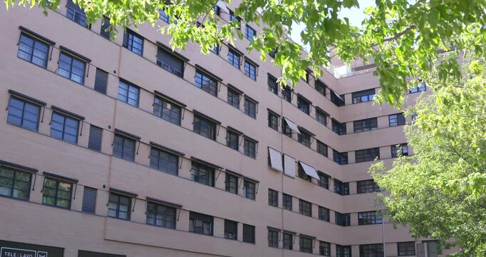Residential apartment building with beige facade and multiple windows facing a public square in Madrid, Spain. Tree branches frame the urban residential architecture under bright daylight.