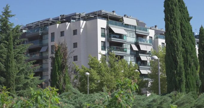 Contemporary residential apartment building with balconies and landscaped gardens in Madrid, Spain. Green trees and shrubs frame the modern housing complex under clear blue sky on a sunny day.