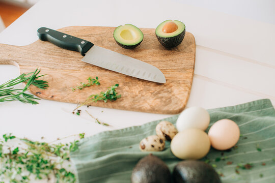 Halved avocado with knife on wooden board, herbs and eggs