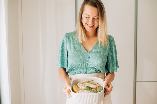 Smiling woman holding raw chicken dish with herbs in home kitche
