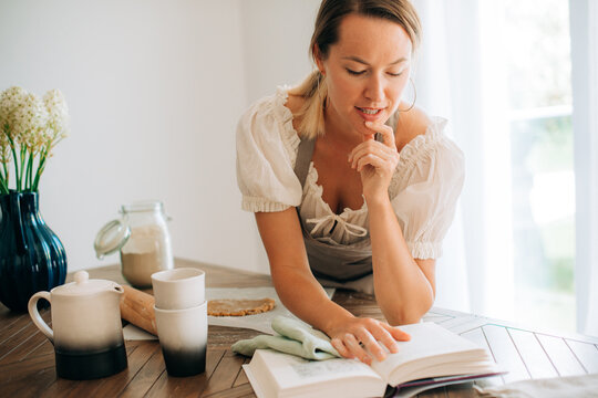 Woman in apron reading recipe book in kitchen