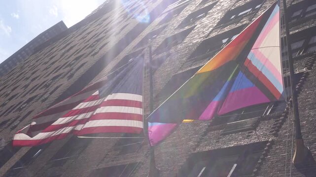 A cinematic video clip of an LGBTQ flag and an American flag waving in front of skyscrapers on a sunny day.
